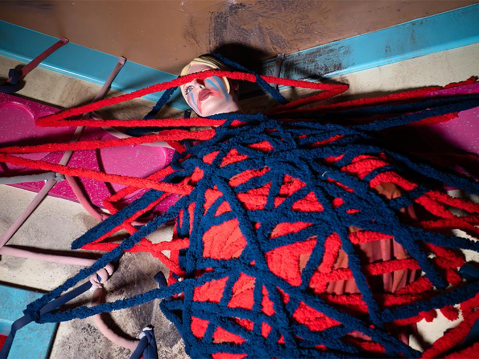 A mannequin doll from Dolls: Let's Play Dead is wrapped tightly in a web of red and blue yarn that crisscrosses over its body and face. The doll has a pale complexion with short blonde hair. Its makeup is dramatic and intentionally smeared with bright blue eyeshadow streaks downward from the eyes like tears. The lips are smeared with vivid red paint. The yarn crosses the face of the doll, covering one eye, adding to the layered, artistic composition. The background features pink heart shapes and teal accents, contributing to the vibrant and surreal atmosphere.