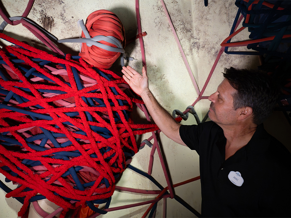 A person stands in front of an elaborate art installation featuring a mannequin wrapped fully in layers of red, blue, and gray oversized yarn pieces. The mannequin is positioned upright against a worn, discolored wall, with additional yarn pieces extending outward in a web-like pattern that covers part of the wall. A person wearing a black shirt with a white Universal Orlando Resort name tag reading “JMAC” raises one hand toward the wrapped figure, appearing to show off the display. The lighting is soft and ambient, highlighting the textures of the fabric and the aged wall behind them. The scene is part of one of the rooms of the haunted house Dolls: Let’s Play Dead, and evokes a sense of eerie creativity through its use of color, material, and spatial design.