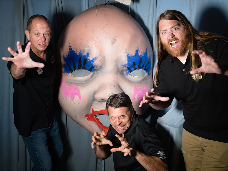 Three people pose playfully in front of a large, eerie baby doll mask installation. The mask features exaggerated makeup, including bright blue eyeshadow, heavy pink blush on the cheeks, and bold red lipstick. Its eyeless pale face and oversized features give it a haunting, theatrical presence. The person on the left, Jamie, wears a black collared shirt and jeans, extending one arm forward with fingers spread wide in a dramatic claw-like gesture. The person in the middle, Jeff, crouches slightly, hands raised like claws, also dressed in a black collared shirt. The person on the right, Chris, has long hair and a full beard, wearing a black collared shirt and khaki pants, mimicking the same claw-like pose with one arm extended toward the camera. Behind them, deep blue curtains frame the scene, adding contrast and depth to the stylized environment. The group stands in one of the rooms of the haunted house Dolls: Let’s Play Dead.