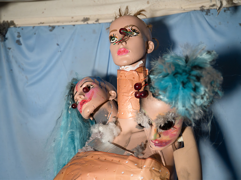 A surreal and eerie art installation features three mannequin doll heads attached to a single torso. The central head is bald with exaggerated makeup including long black eyelashes and pink lipstick. The central head has a pin and wires sticking through its left eye and a neck that is wrapped with what appears to be an adhesive bandage. To the left, a second mannequin head with vibrant blue hair is tilted sideways. It has similar heavy makeup, a pin sticking into each eye and visible neck damage, with stuffed animal stuffing spilling out. On the right, the third head has blue hair styled in a fluffy bun. This head is slightly tilted downward and also features dramatic makeup. The third head has the eyes of a stuffed animal replacing their doll eyes. All three heads share the same torso, which is partially visible and wrapped in adhesive bandages. Behind them is a light blue curtain, stained and worn near the top edge where it meets the ceiling. The scene is part of the haunted house, Dolls: Let’s Play Dead, and evokes a sense of decay, distortion, and theatrical horror through its damaged materials and unnatural composition.