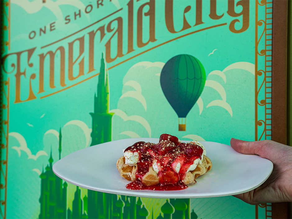 A hand holding a plate with the Strawberriful Cheesecake Waffle in front of an Emerald City poster in Universal Studios Florida.
